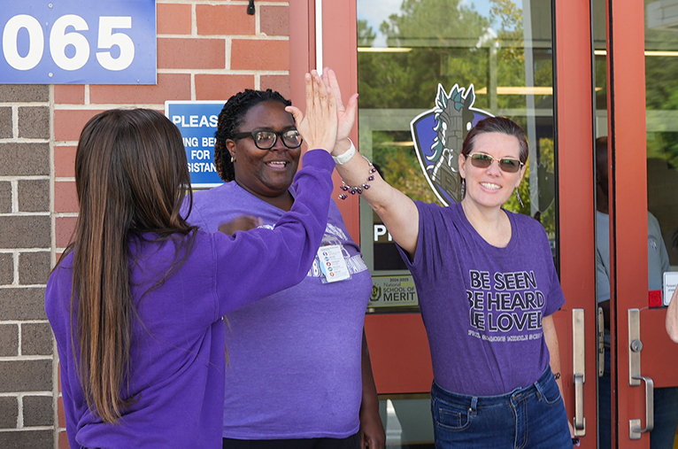 Three women in purple shirts give high fives in front of a building. The building has a brick wall and a sign with number 065.