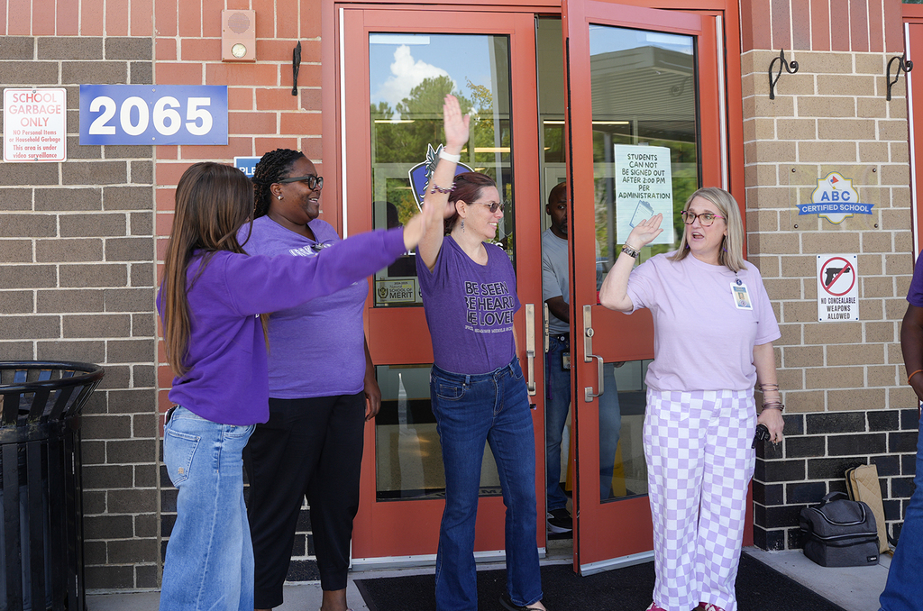 Four women in matching shirts raise their hands in front of a building. A trash can is nearby.