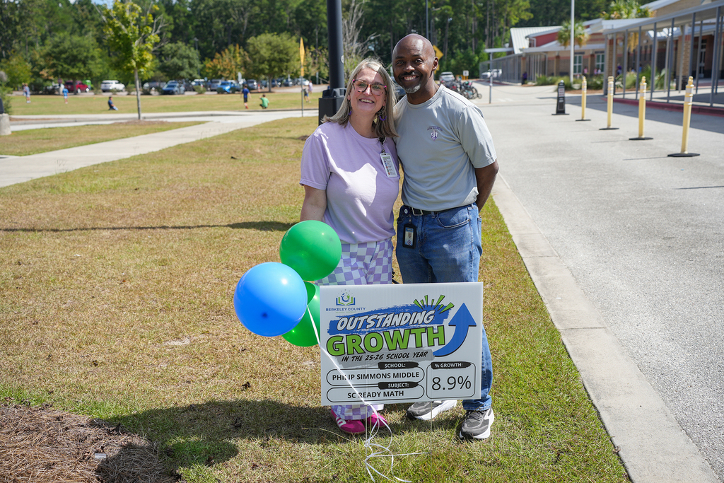 Two people stand by a sign reading "OUTSTANDING GROWTH" with green balloons in a grassy area.