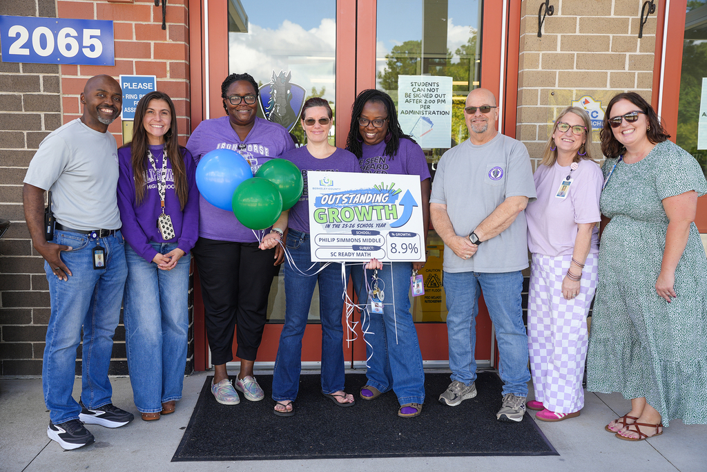Nine people stand in front of a building with balloons. The group holds a sign with text and a percentage.