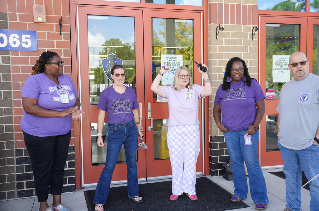 Five people in matching purple t-shirts stand in front of a building. A woman in the middle holds a pair of scissors.