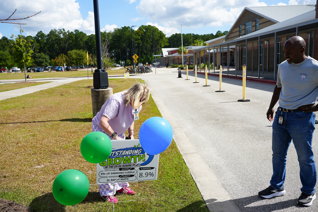 A woman kneels on grass by balloons next to a sign, with a man standing nearby. A road with yellow posts stretches behind them.