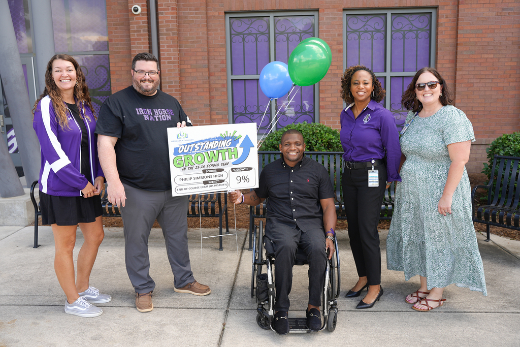 Six individuals stand outside a brick building, two in wheelchairs, holding a large sign with green arrows.