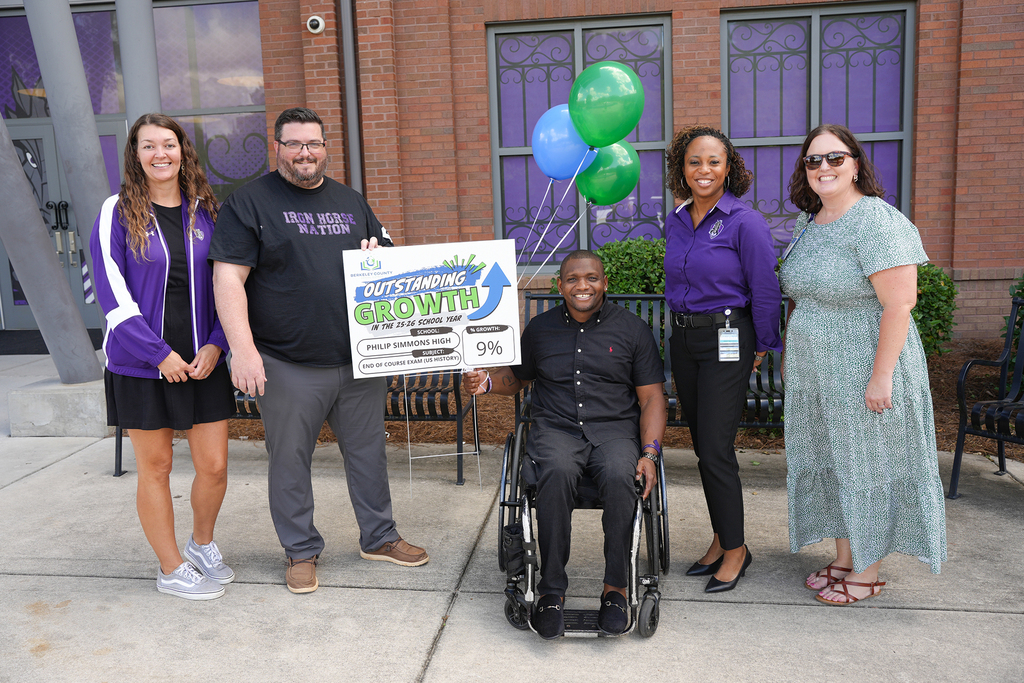 Six people, including a man in a wheelchair, stand on a sidewalk outside a building. They pose for a picture.