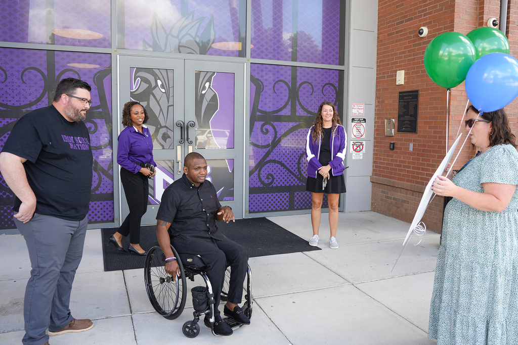 Five people gather in front of a building with a purple and green design. A man in a wheelchair holds balloons.