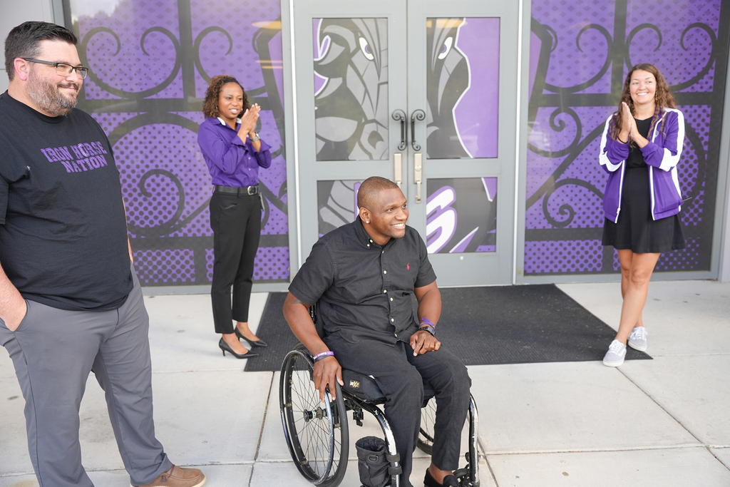 Four people stand outside a building with a purple and black backdrop. A man in a wheelchair smiles at the camera.