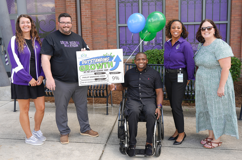 A group of six individuals pose for a photo on a sidewalk, one in a wheelchair. They hold a sign.