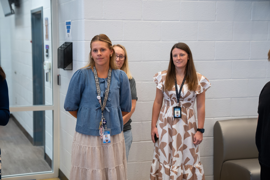Two women stand in a hallway, both wearing IDs and smiling. One wears a denim top, the other a floral dress.