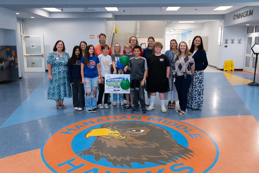 A group of people, including children and adults, stand in a hallway with a school emblem on the floor.
