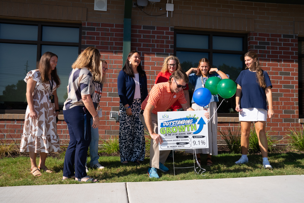A group of people stands outside a brick building. A man bends down, looking at a sign with balloons.