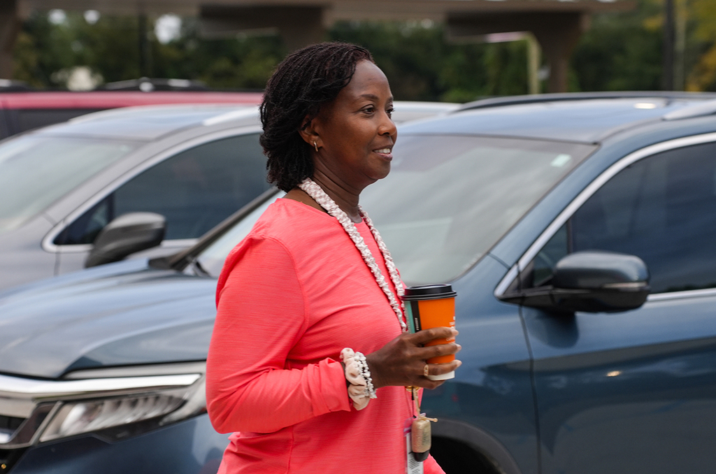 A person in a coral top stands in a parking lot, holding a coffee cup and facing away from two blue cars.