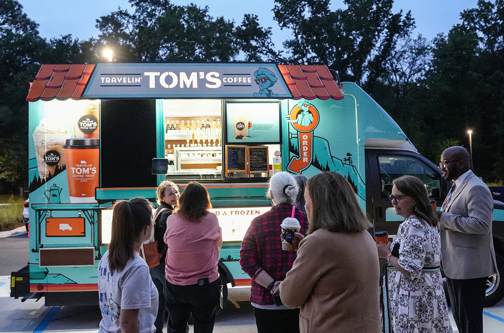 A colorful Tom's Coffee truck stands in a parking lot. People, some holding cups, gather around it.
