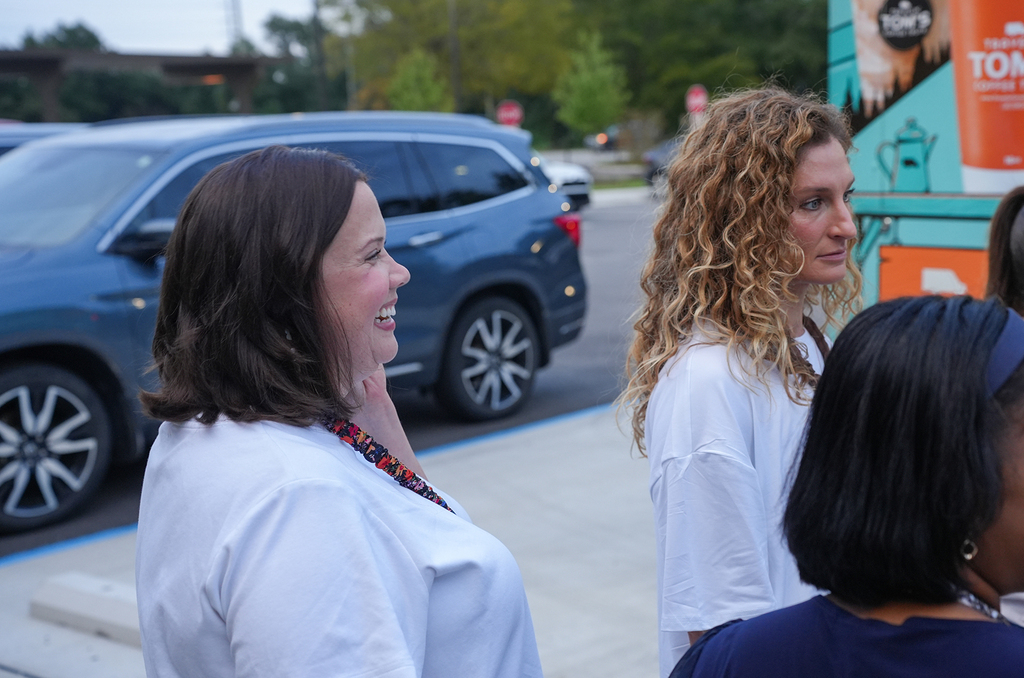 Several women standing outdoors. One wears a necklace. A car is parked on the side of the road.