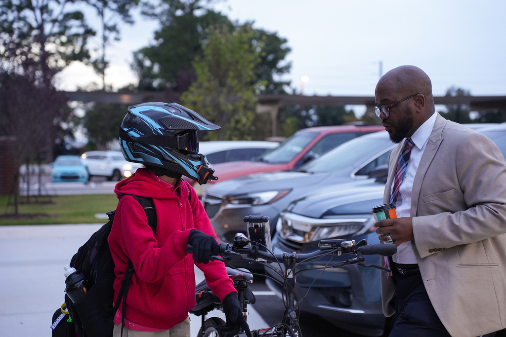 A man in a tan suit speaks to a child on a bicycle. The child wears a helmet and a red hoodie.