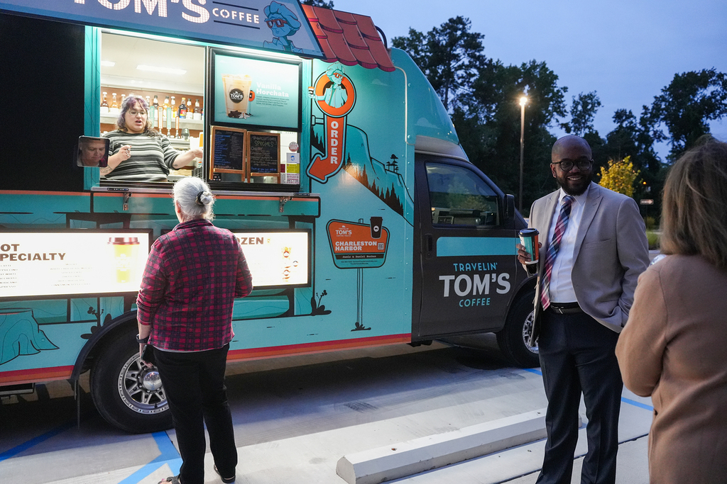 A Tom's Coffee truck is parked, with a barista inside. Two individuals are standing in front of the truck.