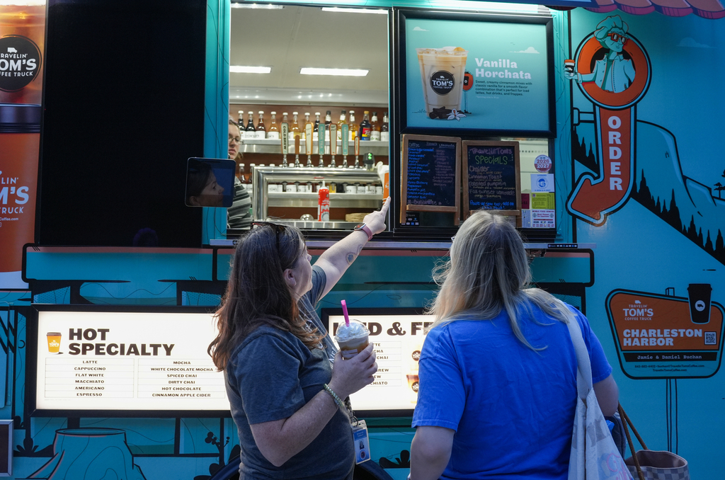 Two women stand at a food truck with a menu board and drinks. The truck has blue and white designs.