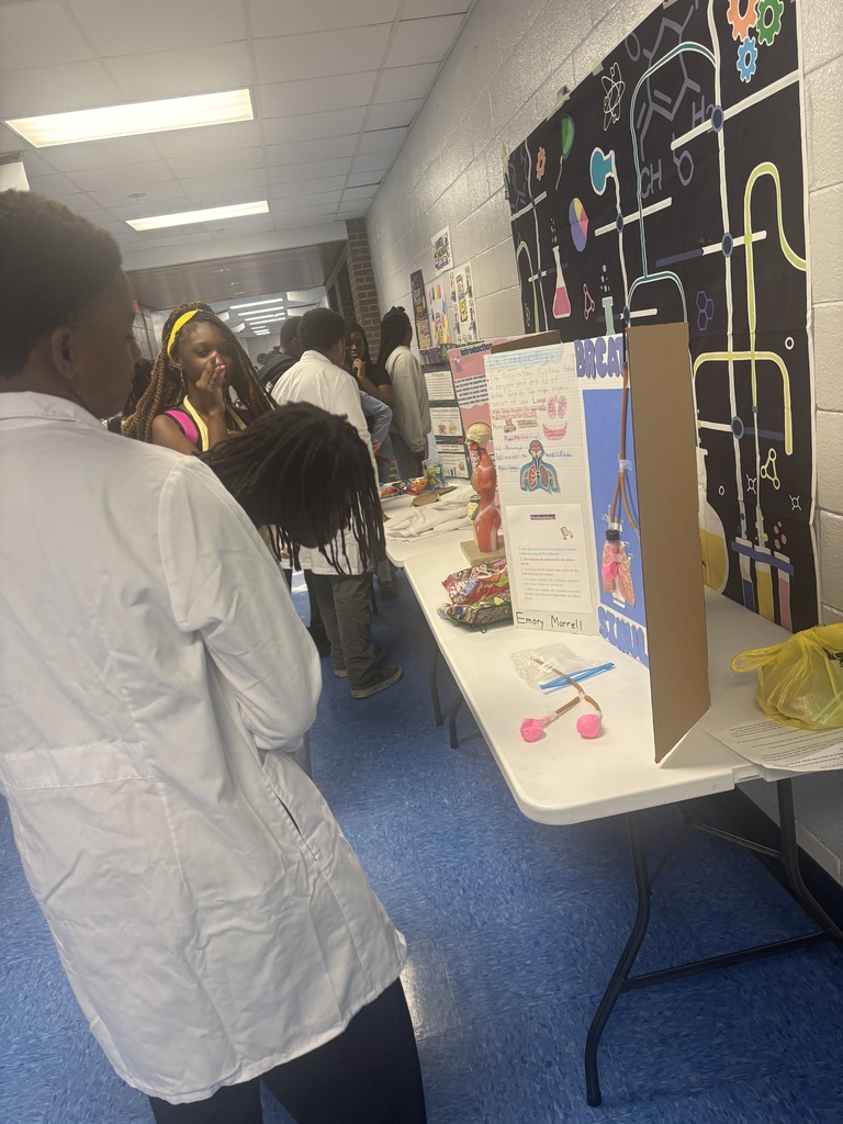 People in white lab coats view a project display at a school event. A table holds a cardboard box, posters, and tools.