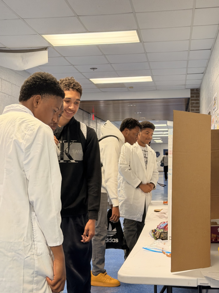 Four students in lab coats in a hallway with a display table, a box, and items.