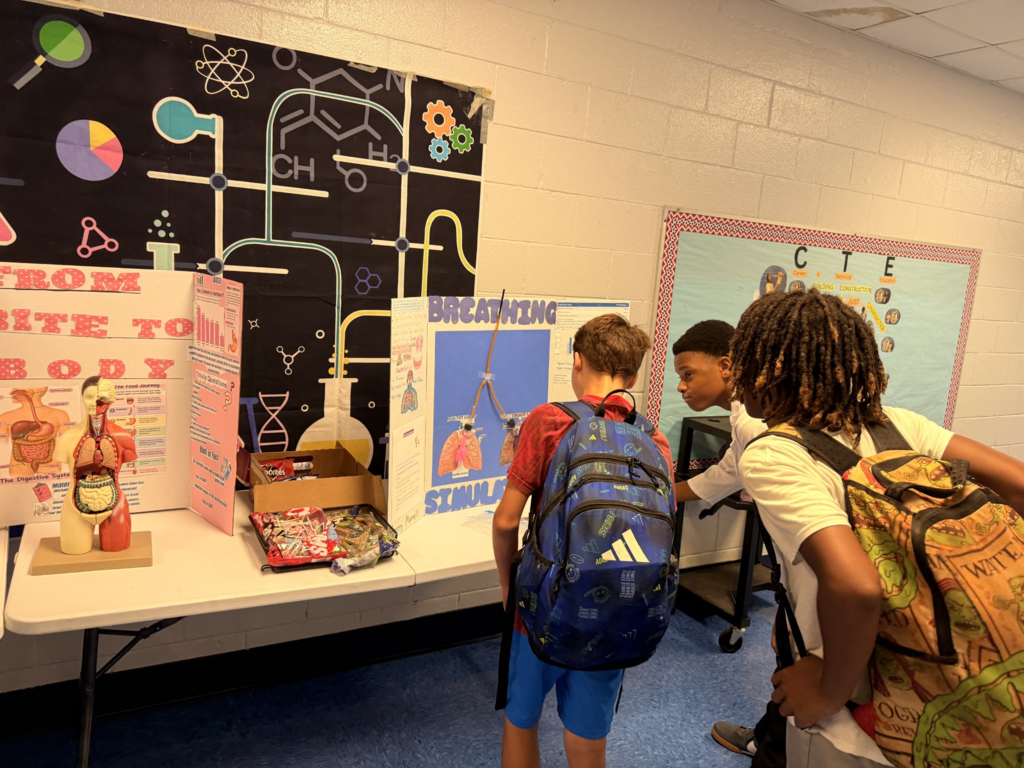 Three kids view a table with a model, posters, and boxes. One has a backpack with a logo.