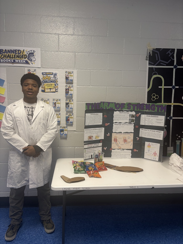 A person in a white lab coat stands next to a white table with a display of objects and papers.