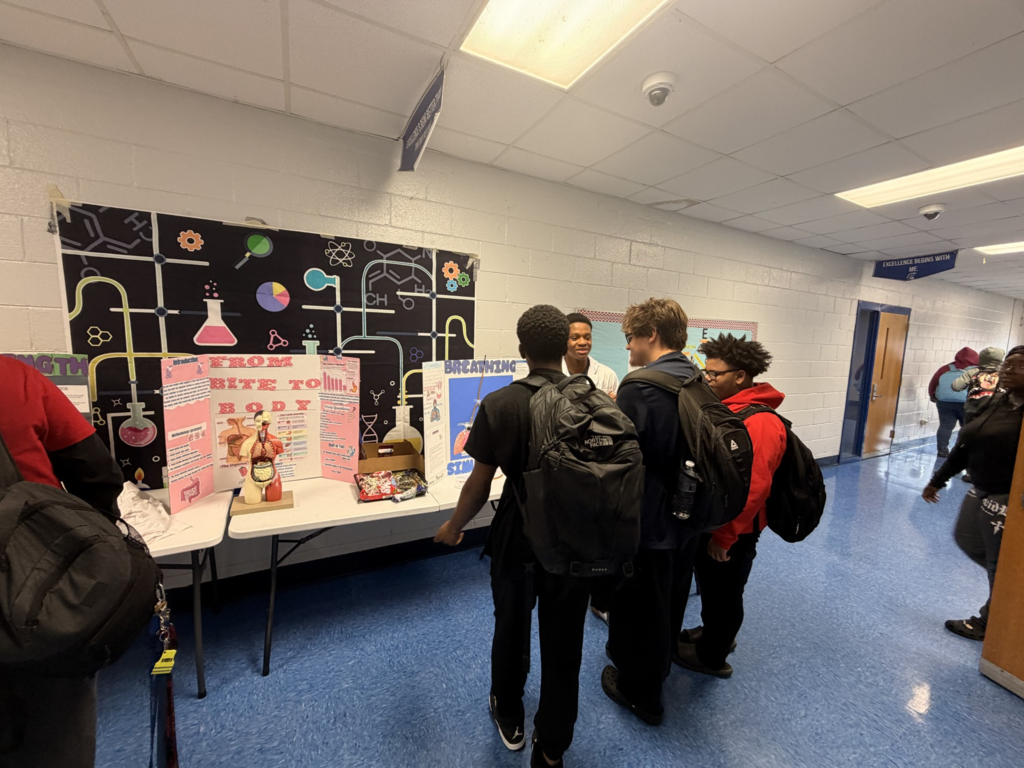 People stand around a table with a science project. The hallway has white walls and blue flooring.