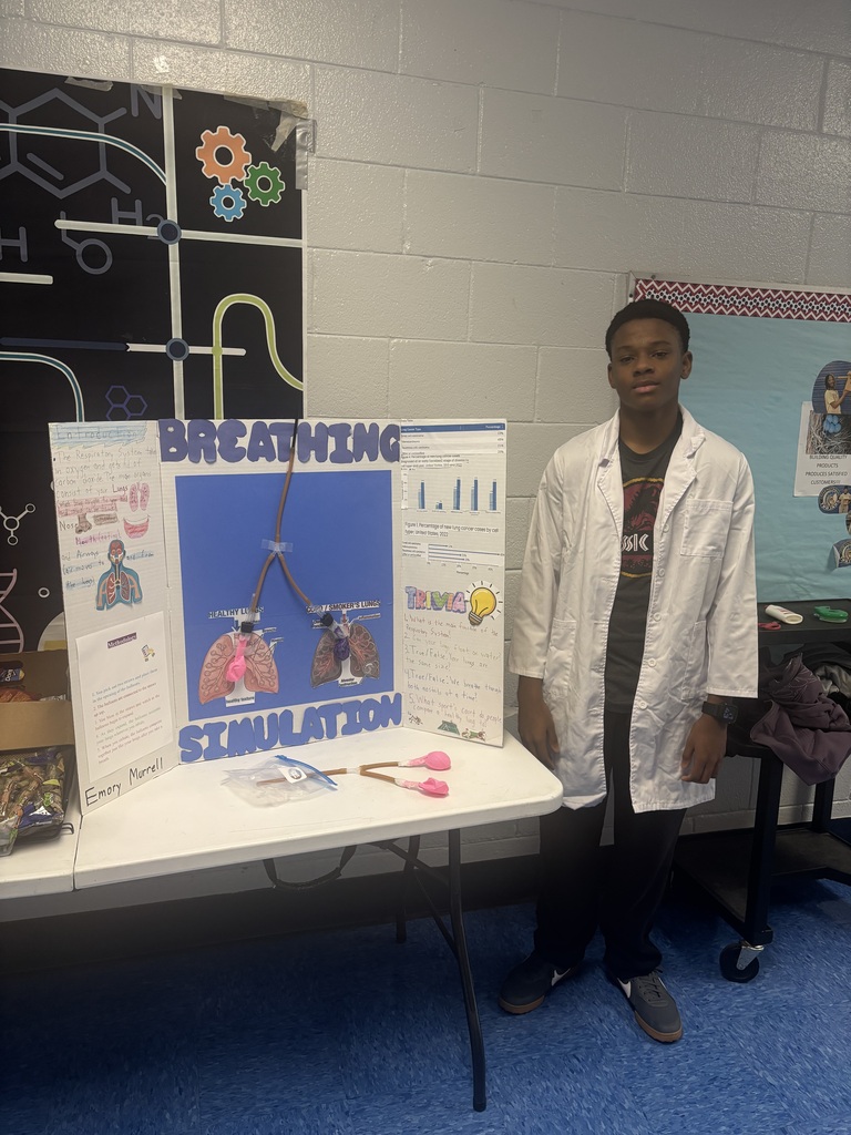 Person in lab coat stands by a table with a sign that reads "Breathing Stimulation" against a white wall.