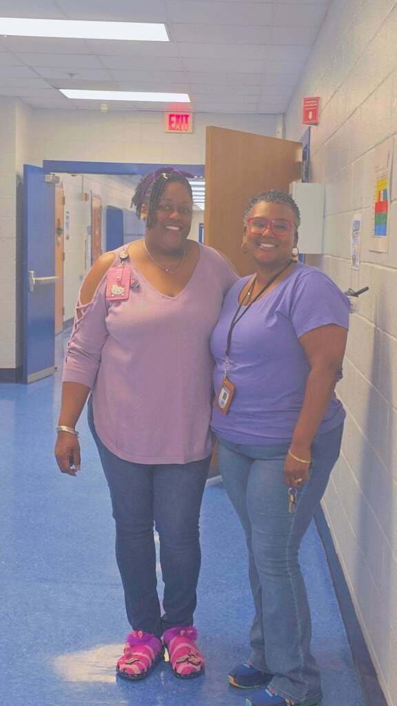 Two people in similar shirts stand close to each other in a hallway with white walls and blue flooring.