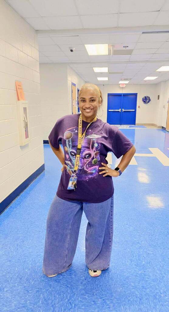 Person with blonde hair wearing a purple t-shirt and blue jeans, standing with hands on hips in a blue hallway.