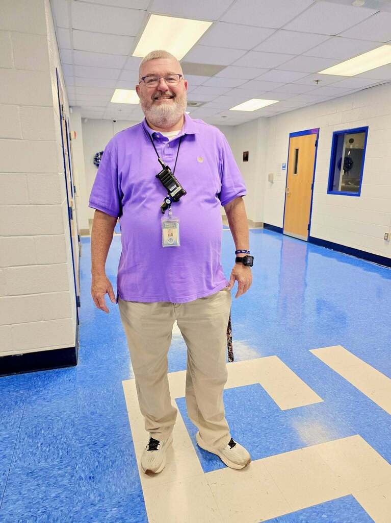 Person in a purple shirt and khaki pants stands in a hallway with blue floors, white walls, and wooden doors.