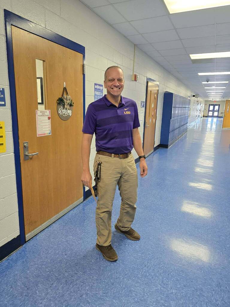 Man in purple shirt and khaki pants standing in hallway with lockers. Floor is blue and walls are white.