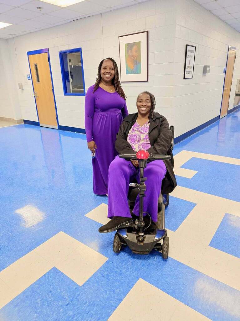 Two women, one in a wheelchair, pose for a picture in a hallway with blue and white flooring.