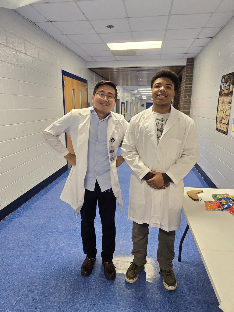 Two men in white lab coats, one with glasses, stand in a brightly lit hallway with blue flooring.