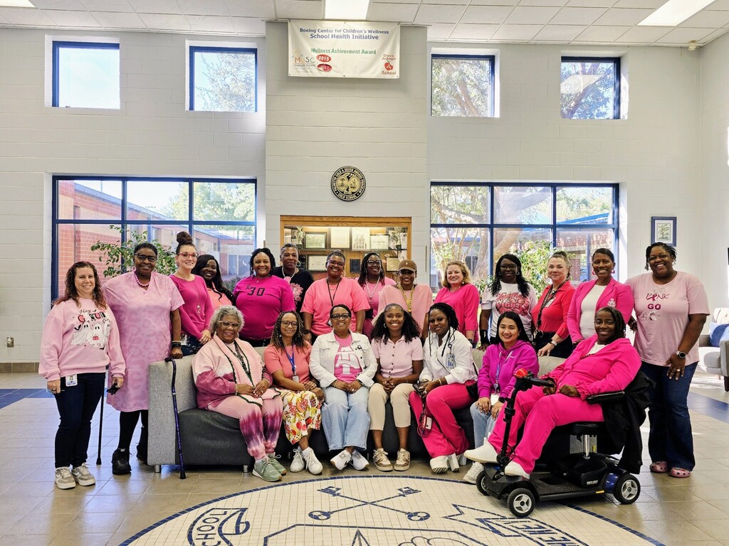 Group of adults in pink shirts and various clothing poses for photo in a room. A rug sits in the center.