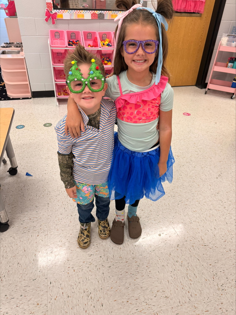 Two children stand close, one wearing glasses and a shirt, the other with a pink top and tutu.