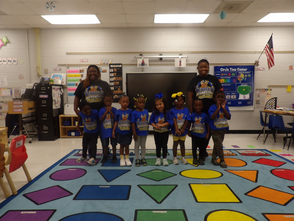 A group of children in blue shirts stand in a classroom. They are surrounded by educational materials and a patterned rug.