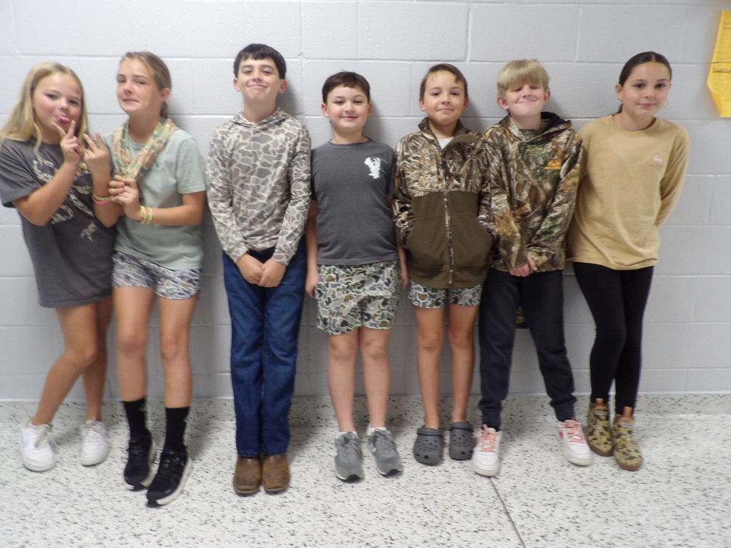 Six children in matching clothing stand together against a plain wall. They are wearing jackets and shorts.