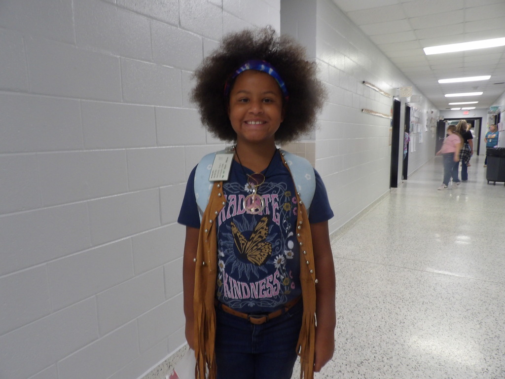 A smiling child with an afro stands in a school hallway, wearing a blue shirt and brown vest.