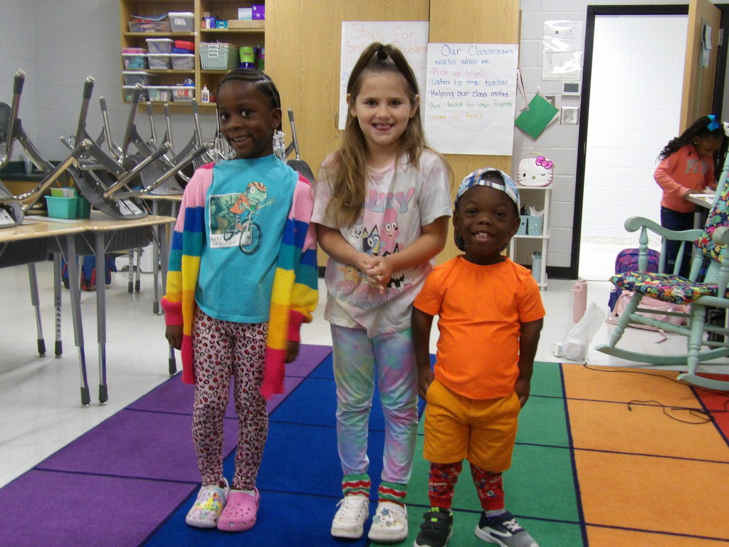 Three children stand on a multi-colored floor mat, smiling. A classroom with desks, chairs, and a rocking chair is in the background.