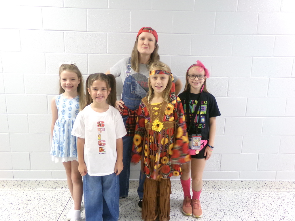 Five children and an adult woman stand in a hallway. The woman wears a red headband and denim overalls. The children wear diverse outfits.
