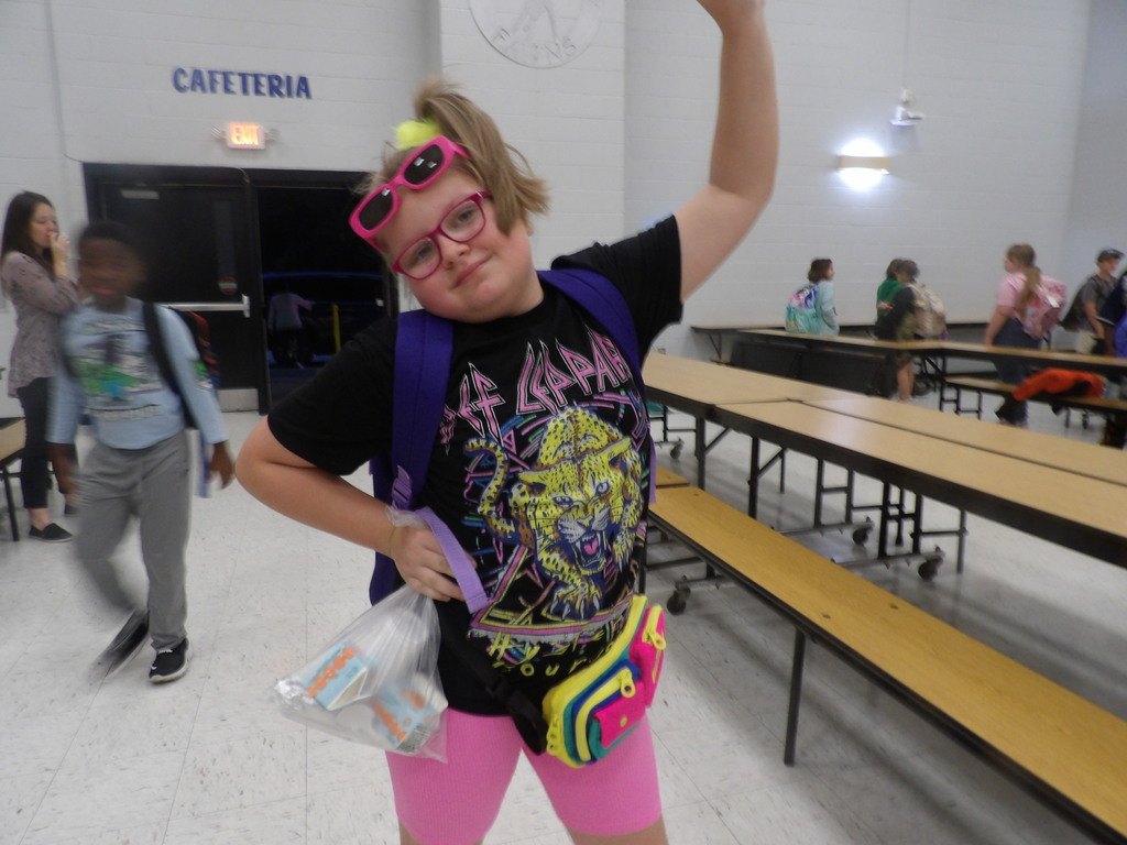 A child wearing a black t-shirt, pink glasses, and a pink backpack stands with one arm raised.