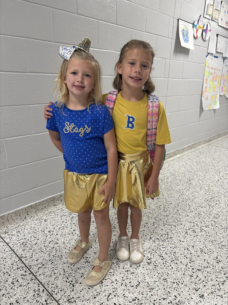 Two young girls wearing gold shorts and white shoes. One has a blue shirt with white dots, and the other has a yellow shirt with a B.