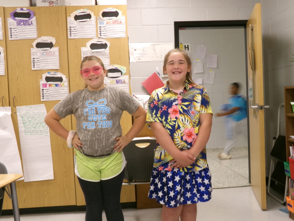 Two girls stand in a classroom. One wears glasses and a gray T-shirt; the other wears a floral dress. A door is open behind them.