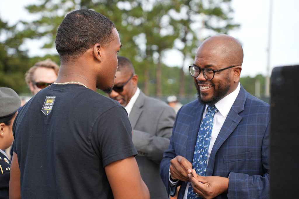 Two men speak to each other in front of a blurry background of trees and other people.