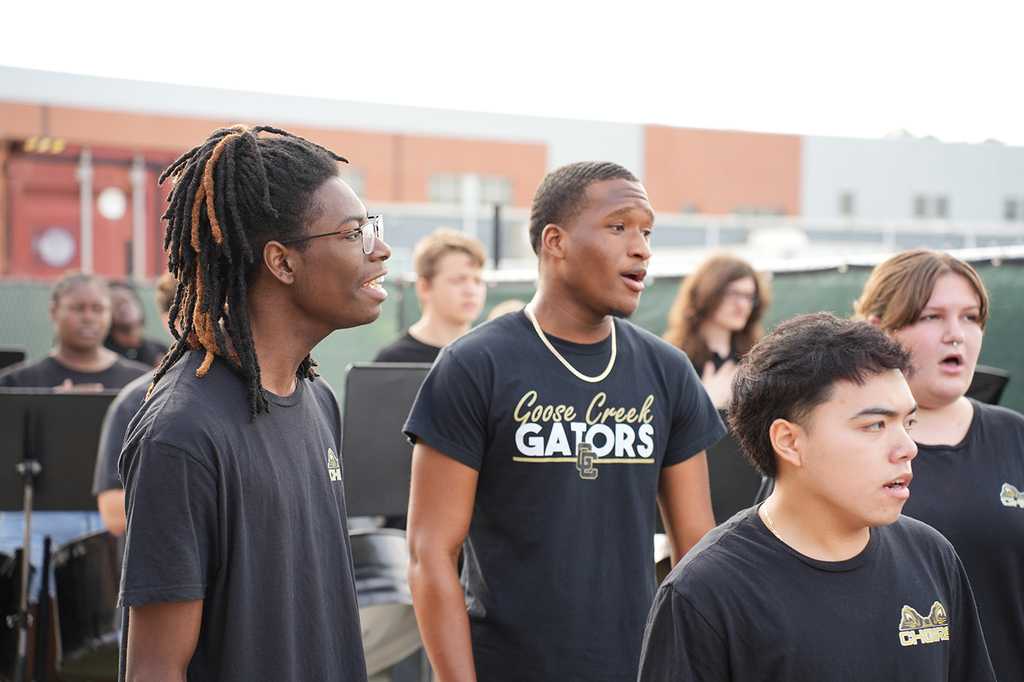 Three people in black shirts with the word "Gators" sing with others. Behind them, several people are seated.