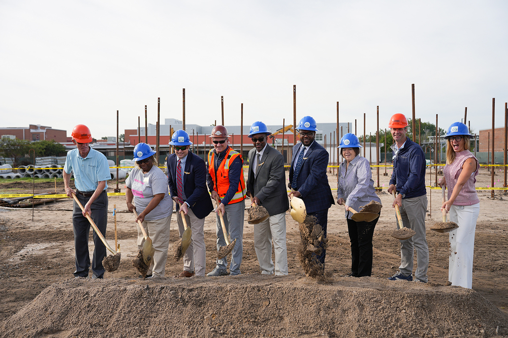 A group of people at a construction site, all wearing hard hats, holding shovels, and standing on a dirt mound.