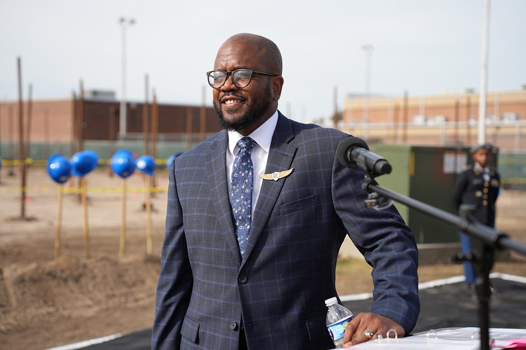 A man in a suit stands at a podium, smiling. Behind him, a field with construction, blue hard hats, and a building.
