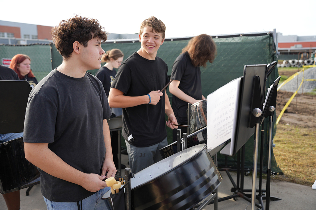 Four people play drums outside. A young man in the foreground holds drumsticks. The background is a green fence.