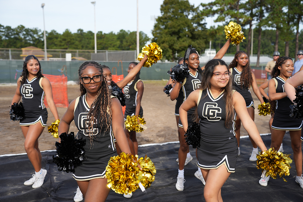 Girls wearing black and gold cheerleading uniforms perform on a field. Background includes trees and a fence.
