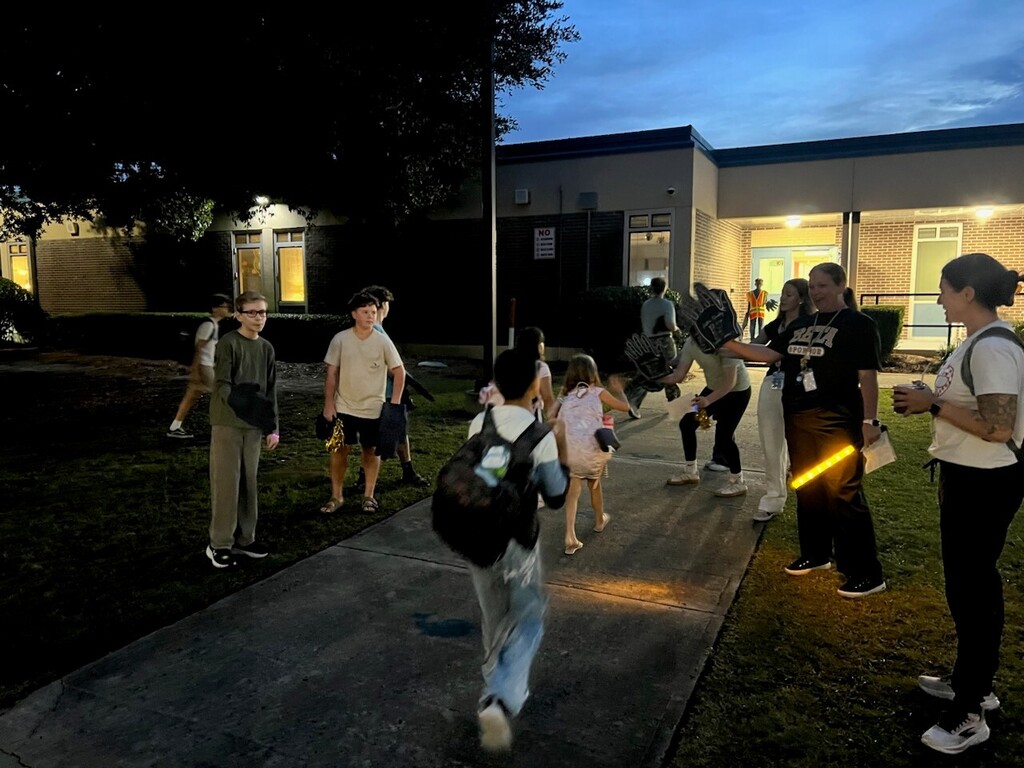 People, including children, walk on a sidewalk at night, illuminated by streetlights. Some carry bags.