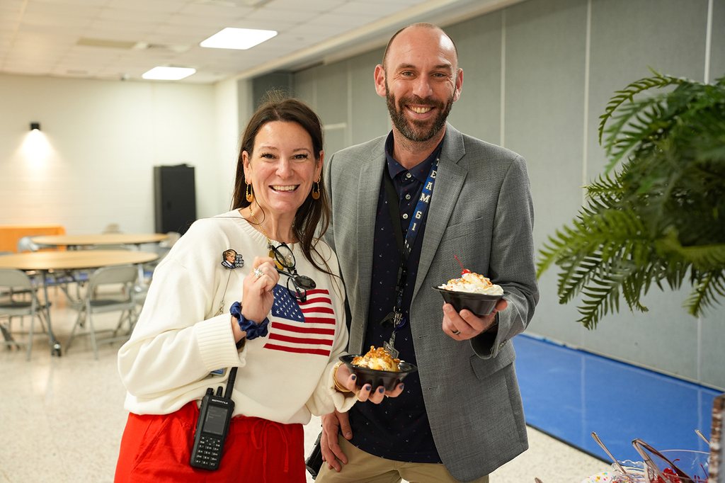A man and a woman smiling, holding bowls of ice cream, standing in a room with tables, chairs, and a potted plant.